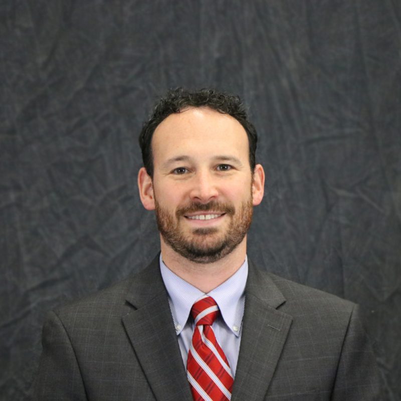 Photo of Bill Melleby, he has short brown hair, short beard, wearing a gray suit jacket, blue shirt, and red tie.
