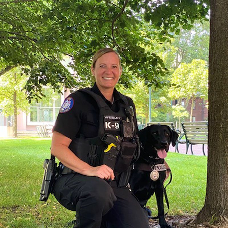 Police Officer Wesley, wearing a black police uniform, poses with canine Uman, a black labrador retriever, in a field under a tree.