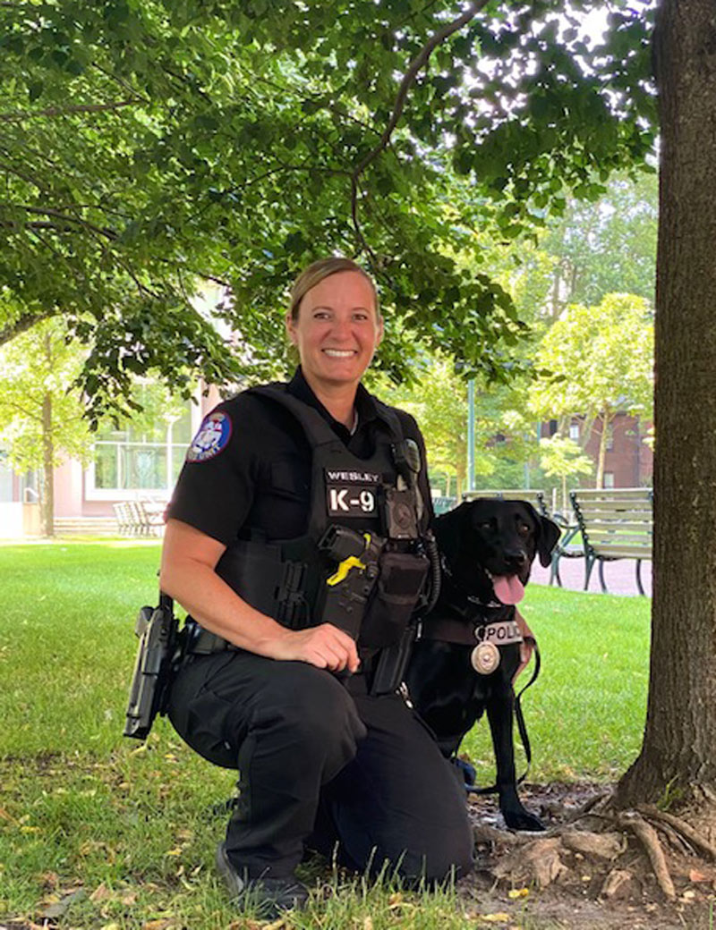 Police Officer Wesley, wearing a black police uniform, poses with canine Uman, a black labrador retriever, in a field under a tree.